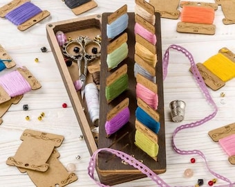 May include: A wooden box with a variety of colorful threads and embroidery floss spools, a pair of scissors, and a thimble. The box is open and the threads are arranged in a neat row. There is also a spool of lace and a few beads scattered around the box.