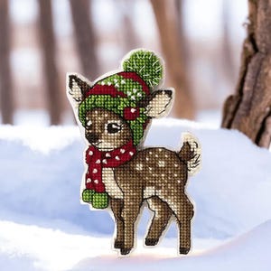 May include: A brown and white spotted fawn wearing a green and red knitted hat with a white pom pom and a red and white polka dot scarf. The fawn is standing in the snow.