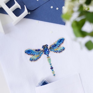 May include: A white shirt with a blue and green dragonfly embroidered on the front. The dragonfly is made of beads and sequins. The shirt has a pocket on the front.