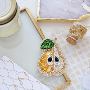May include: A beaded pear brooch with a white, yellow, and green color scheme. The pear is made of white and gold beads, with yellow beads forming the pear's shape. The pear has a green leaf made of green beads.