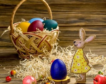 Può includere: Un uovo di Pasqua blu si trova in un porta uova di legno a forma di coniglio. Il coniglio è decorato con perline gialle e bianche. Un cestino di vimini pieno di uova di Pasqua colorate si trova sullo sfondo.