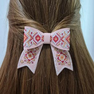 May include: A light pink hair bow with a cross-stitch design. The bow features a repeating pattern of red, orange, and yellow floral motifs on a white background. The bow is attached to a hair clip.