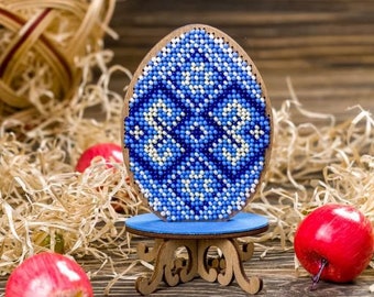 Può includere: Un ornamento a forma di uovo in perline blu e bianche su un supporto in legno. L'uovo è decorato con un motivo geometrico ed è esposto su un supporto in legno con intagli intricati.