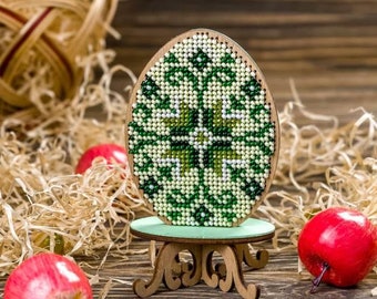 Può includere: Un designo di uovo di Pasqua con perline verdi e bianche su un supporto in legno. L'uovo è decorato con un motivo geometrico.