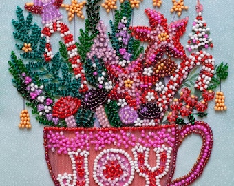 Puede incluir: Una pieza de arte bordada con cuentas que muestra una taza rosa con la palabra JOY escrita en cuentas blancas. La taza está llena de un arreglo festivo de ramas de pino verdes, bastones de caramelo rojos y blancos, estrellas y un árbol.