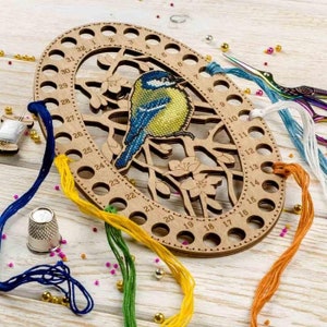 Può includere: Un organizzatore di fili in legno con 32 fori per riporre il filo da ricamo. L'organizzatore ha la forma di un uccello su un ramo con un design blu e giallo. L'organizzatore è circondato da filo da ricamo colorato.