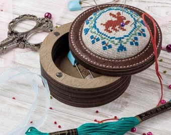 May include: A wooden pincushion with a brown leather top and a circular embroidered design of a rabbit in red, blue, and green. The pincushion is open and has a magnetic closure. There are several pins stuck in the pincushion. A spool of teal embroidery floss is in the foreground.