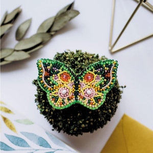 Può includere: Una spilla a forma di farfalla con perline verdi, gialle, arancioni, rosa e bianche. La spilla è appoggiata su un letto di muschio verde.