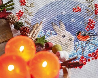 Könnte beinhalten: Ein weißer bestickter Tischläufer mit einer Winterszene mit einem weißen Kaninchen und einem Rotkehlchen. Die Szene ist von roten Beeren und einem blauen Hintergrund umgeben. Drei rote Kerzen befinden sich im Vordergrund.