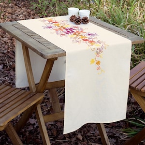May include: A white table runner with an embroidered design of autumn leaves in shades of orange, yellow, and red. The runner is on a wooden table with two chairs.