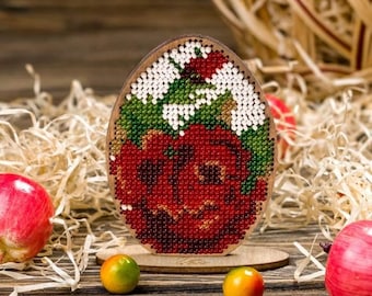 Può includere: Un ornamento di legno a forma di uovo con un motivo di perline a forma di rosa rossa e foglie verdi. L'ornamento è su un supporto di legno.