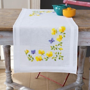 Peut inclure: Un chemin de table blanc avec des fleurs jaunes et des papillons violets brodés. Le chemin de table est sur une table en bois avec un piètement peint en blanc.