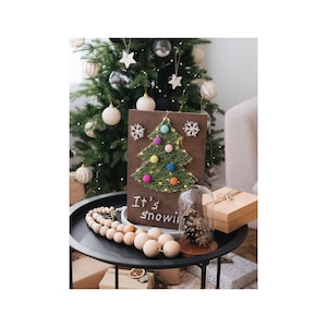 Peut inclure: Une œuvre d'art de Noël en fil sur un fond en bois brun avec un arbre de Noël en fil et des pompons colorés. Le texte "It's snowing" est écrit en dessous.