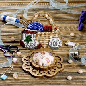 Pode incluir: Um almofadinha para alfinetes de madeira com um design floral decorativo. O almofadinha para alfinetes tem uma tampa removível que revela um pequeno compartimento para guardar agulhas de costura. O almofadinha para alfinetes é decorado com uma imagem de contas de uma cesta cheia de fio e agulhas de tricô.