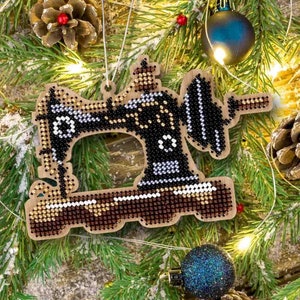May include: A wooden Christmas bauble shaped like a sewing machine, decorated with black, white and gold beads. The bauble is hanging on a green pine branch.