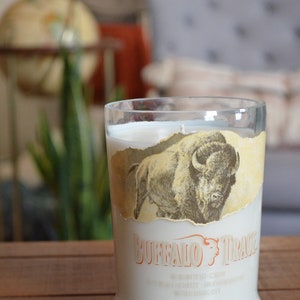 May include: A clear glass candle jar with a white candle inside. The jar has a label with a drawing of a buffalo and the text "Buffalo Trace" in orange. The label also includes the text "Kentucky Straight Bourbon Whiskey" in white.