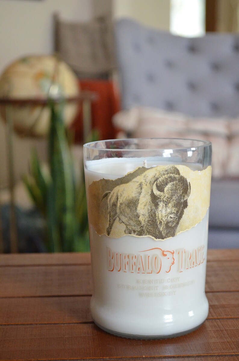 May include: A clear glass candle jar with a white candle inside. The jar has a label with a drawing of a buffalo and the text "Buffalo Trace" in orange. The label also includes the text "Kentucky Straight Bourbon Whiskey" in white.