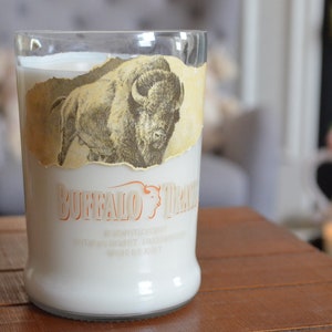 May include: A white candle in a clear glass bottle with a vintage label featuring a buffalo illustration and the text "Buffalo Trace" in orange. The label is torn and faded.