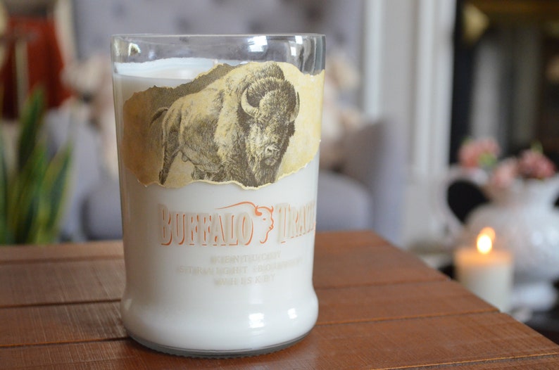 May include: A white candle in a clear glass bottle with a vintage label featuring a buffalo illustration and the text "Buffalo Trace" in orange. The label is torn and faded.