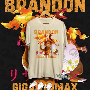 Puede incluir: Camiseta color crema con un diseño de dibujos animados de Charizard, llamas y texto japonés. La palabra "BRANDON" está impresa encima del diseño. El fondo incluye llamas naranjas y amarillas y una gran ilustración de Charizard.