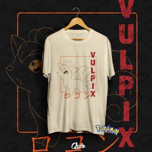 Peut inclure: T-shirt crème avec un motif Vulpix dans un cadre rectangulaire rouge. Le mot "Vulpix" est écrit verticalement en rouge, avec des caractères japonais en dessous. Le t-shirt est suspendu à un cintre en bois.