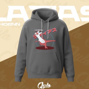 Puede incluir: Una sudadera con capucha gris oscuro con bolsillo de canguro y cord&oacute;n. La parte delantera presenta un gr&aacute;fico de una criatura roja y blanca con el texto "HOENN" encima y "LATIAS" en letras blancas grandes. El logotipo de Qylo est&aacute; en la parte inferior.