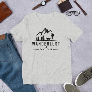 May include: A heather grey t-shirt with a black graphic of a person and a dog hiking in the mountains. The text "WANDERLUST & DOGS" is printed below the graphic.
