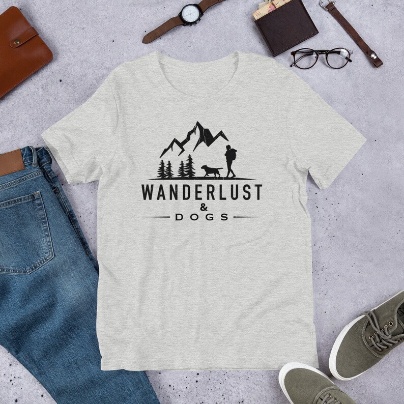 May include: A heather grey t-shirt with a black graphic of a person and a dog hiking in the mountains. The text "WANDERLUST & DOGS" is printed below the graphic.
