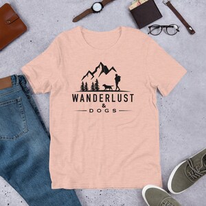 May include: A light pink t-shirt with a black graphic design of a person and a dog hiking in the mountains. The text "WANDERLUST & DOGS" is printed below the graphic.