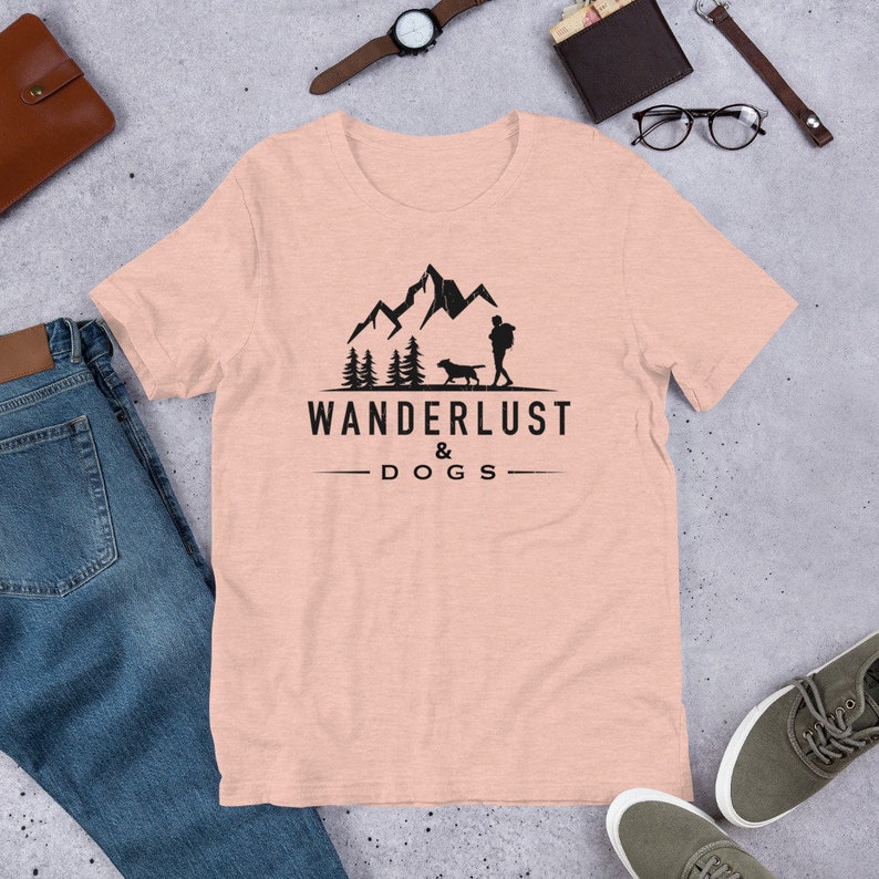 May include: A light pink t-shirt with a black graphic design of a person and a dog hiking in the mountains. The text "WANDERLUST & DOGS" is printed below the graphic.