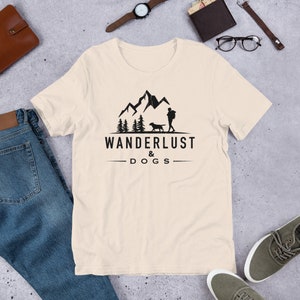 May include: A beige t-shirt with a black graphic design of a person and a dog hiking in the mountains. The text "WANDERLUST & DOGS" is printed below the graphic.