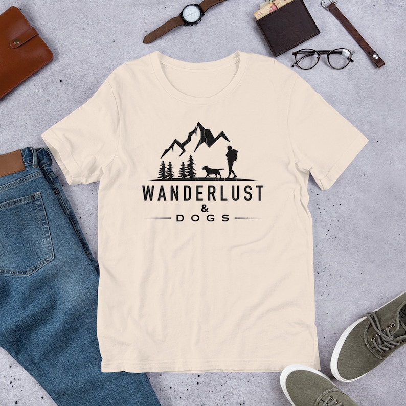 May include: A beige t-shirt with a black graphic design of a person and a dog hiking in the mountains. The text "WANDERLUST & DOGS" is printed below the graphic.