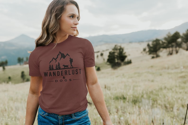 May include: A maroon t-shirt with a graphic of mountains, a person hiking, and a dog. The text "Wanderlust & Dogs" is printed below the graphic.