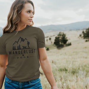 May include: A woman wearing a green t-shirt with a black graphic of mountains, trees, a person, and a dog. The graphic also includes the text "Wanderlust & Dogs".