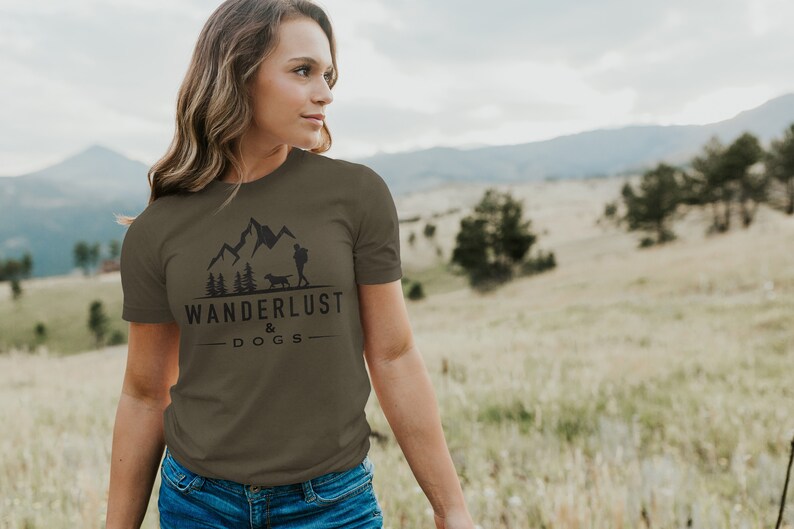 May include: A woman wearing a green t-shirt with a black graphic of mountains, trees, a person, and a dog. The graphic also includes the text "Wanderlust & Dogs".