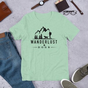 May include: A light green t-shirt with a black graphic of a person and a dog hiking in the mountains. The text "Wanderlust & Dogs" is printed below the graphic.