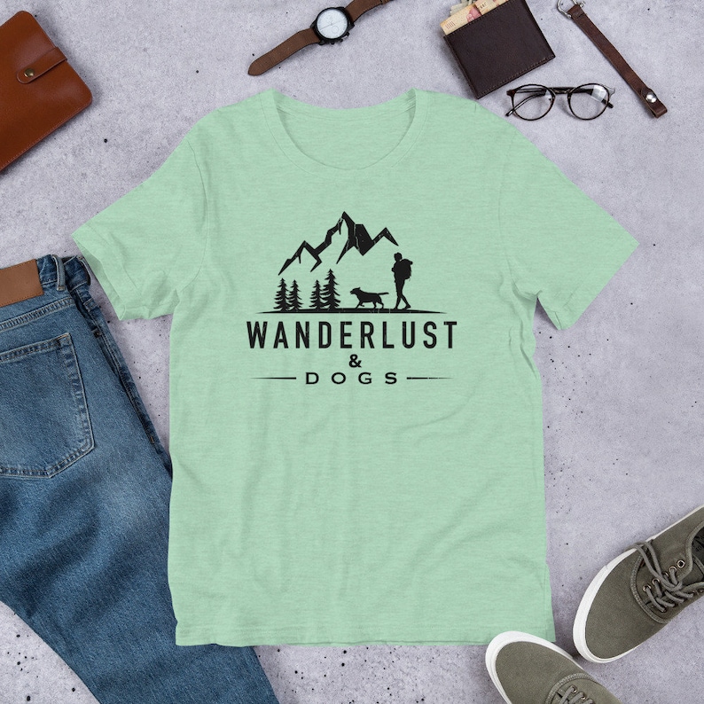 May include: A light green t-shirt with a black graphic of a person and a dog hiking in the mountains. The text "Wanderlust & Dogs" is printed below the graphic.