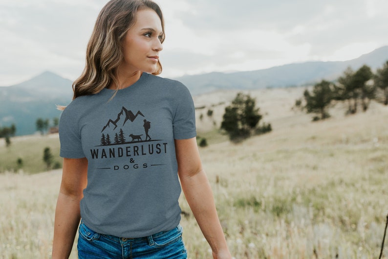 May include: A woman wearing a blue t-shirt with a graphic of mountains, trees, a person, and a dog. The text on the shirt reads "Wanderlust & Dogs".