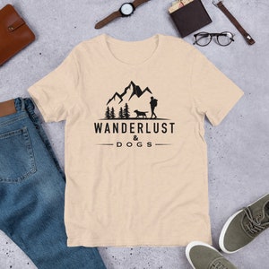 May include: A beige t-shirt with a black graphic of a person and a dog walking in front of mountains and trees. The text "WANDERLUST & DOGS" is printed below the graphic.