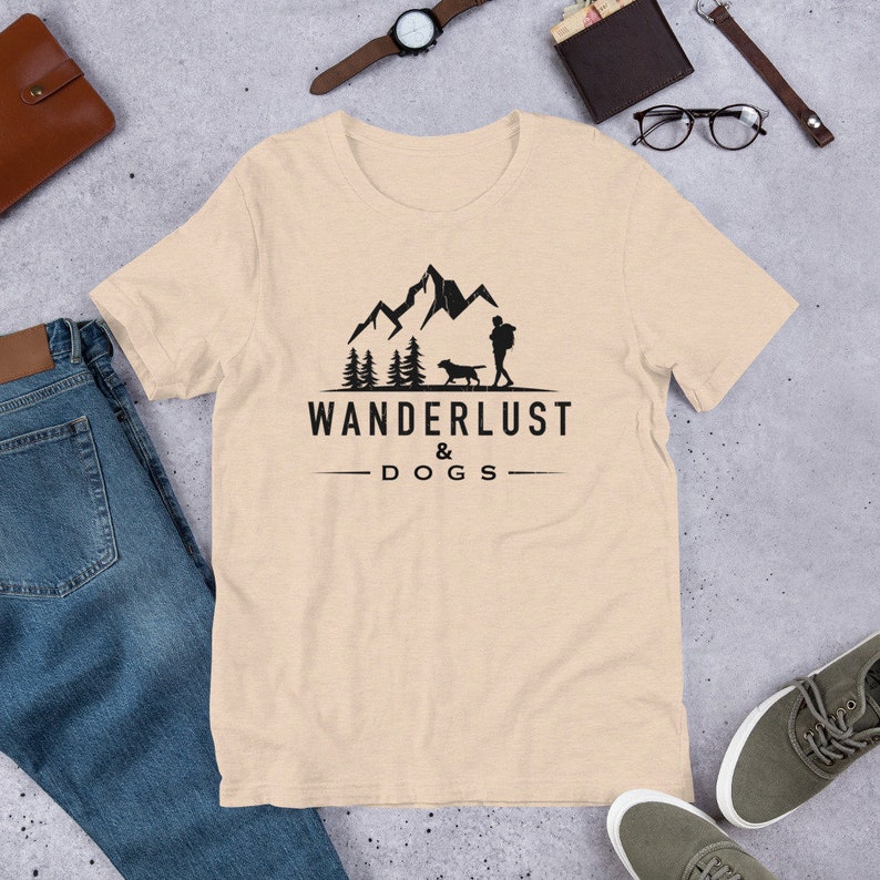 May include: A beige t-shirt with a black graphic of a person and a dog walking in front of mountains and trees. The text "WANDERLUST & DOGS" is printed below the graphic.