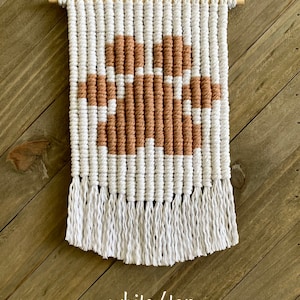 Può includere: Arazzo da parete in macramè bianco e beige con un motivo a forma di zampa e frange.