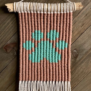 Pode incluir: Uma tapeçaria de parede feita de fio tecido em cores marrom e turquesa. A tapeçaria tem uma franja na parte inferior e está presa a uma vareta de madeira.