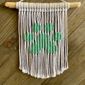 Pode incluir: Uma tapeçaria de macramé branca e verde-água com um design de pegada de pata. A tapeçaria está presa a uma vareta de madeira.