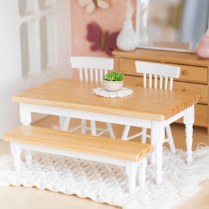 May include: A miniature white table and bench set with a wooden top and white painted legs. The table has a small green plant in a white pot on top of a white crocheted doily.