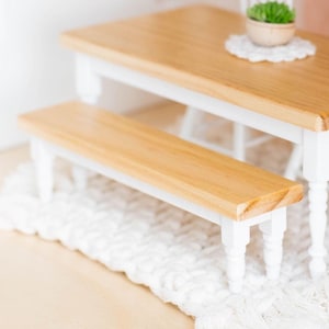 May include: A miniature wooden dining set with a light wood tabletop and white legs. The set includes a table, bench, and chair, with a small plant on the table. The set is placed on a white, textured rug.