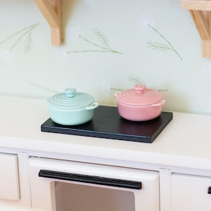 May include: Miniature kitchen scene with a white countertop, featuring a black cooktop holding a light blue and a pink lidded pot. A wooden range hood and shelves with a vase and jars are visible.