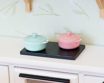 May include: Miniature kitchen scene with a white countertop, featuring a black cooktop holding a light blue and a pink lidded pot. A wooden range hood and shelves with a vase and jars are visible.