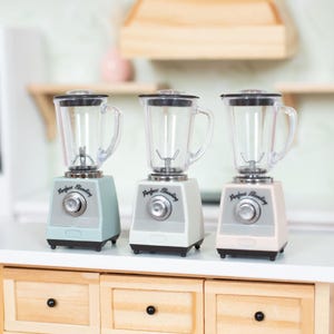 May include: Three miniature blenders in pastel colors: blue, white, and pink. Each blender has a clear pitcher and a silver base with a dial. They are arranged on a white countertop above a wooden cabinet with six drawers and black knobs. The blenders are labeled "Perfect Blending".