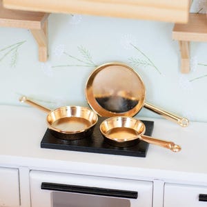 May include: Miniature gold-colored frying pans and a larger pan rest on a black surface, set against a white cabinet. The pans have long handles and are displayed in a kitchen setting. A wooden shelf and a pink vase are also visible.