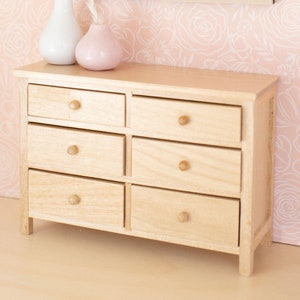 May include: A miniature wooden dresser with six drawers and round knobs. Two framed pictures hang above the dresser, and two vases with dried flowers sit on top. The background features a pink floral wallpaper.